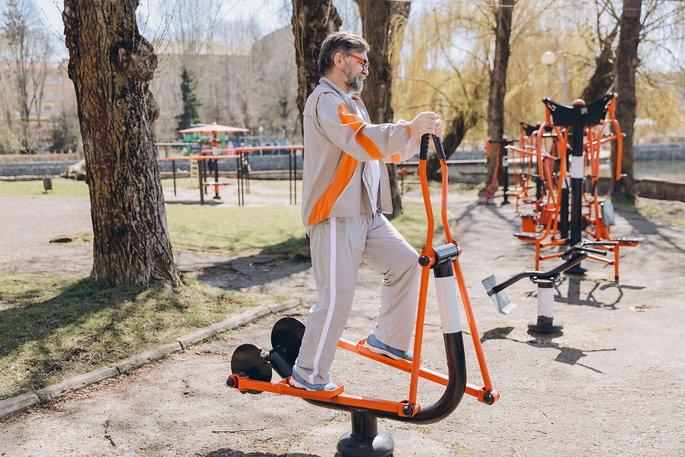 Älterer Mann trainiert im Park auf orangem Outdoor-Trainer. Er trägt grauen Anzug mit orangefarbenen Streifen. Bäume und Geräte im Hintergrund.
