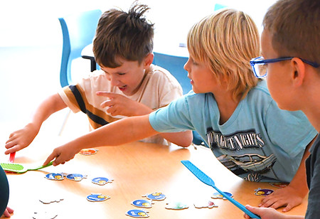 Three kids playing a game on a table with cutouts of bugs and fly swatters.