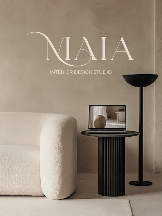 A sophisticated, neutral-toned interior design mockup. The logo "MAIA Interior Design Studio" in elegant white script overlays the image. The scene features a plush, off-white, textured sofa fragment, a fluted black cylindrical side table, and a unique black floor lamp. A silver laptop on the table shows a minimalistic interior on its screen, reinforcing the brand's aesthetic.