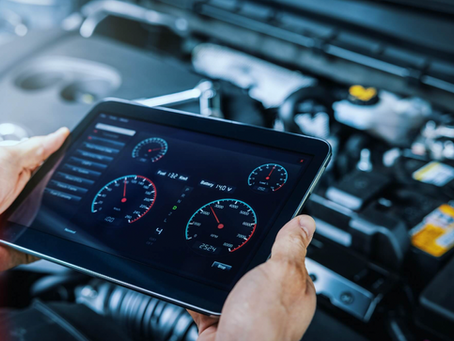 Un écran tactile montrant des jauges numériques de diagnostic de véhicule, tenu par une personne, avec un moteur de voiture en arrière-plan.