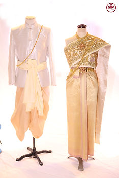 Gold Royal Grace Thai bridal outfit with hand-embroidered gold threads, gemstone belt, and luxurious shoulder drape; groom in ivory silk with pleated pants, golden sash, and ceremonial chain – traditional Thai temple wedding attire.