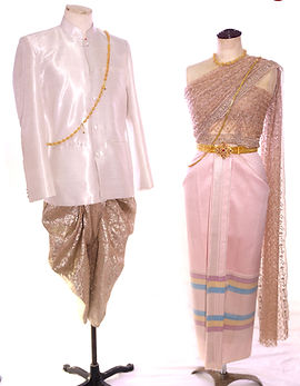Bride wearing pastel rose Chakri Thai traditional dress with rose-gold beadwork and shoulder drape, paired with groom in ivory silk suit and bronze-gold chongkaben trousers, styled for garden or temple wedding