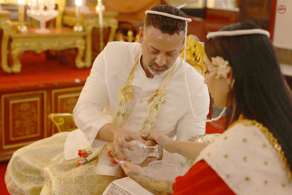 A couple participates in a traditional wedding ceremony, adorned in ceremonial attire and accessories, as they perform a ritual with a crystal vessel.