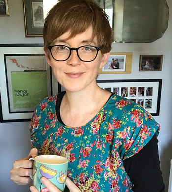 A woman with red hair and glasses is smiling. She holds a mug in her hand and stands in front of a gallery wall of photographs and artwork
