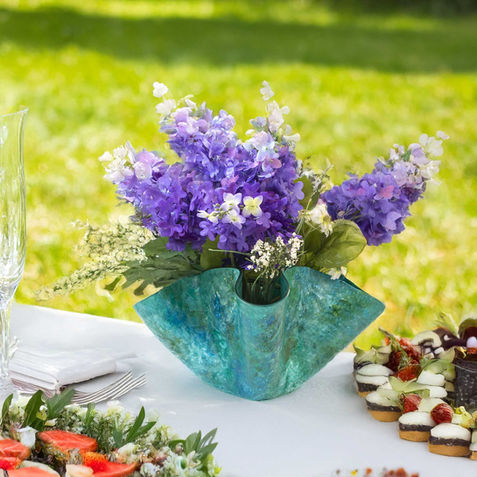 Handmade Emerald Wave Vase in rich green glass with a flowing, wave-like shape. Displayed on a white tablecloth at an outdoor picnic, filled with purple and white flowers, surrounded by fresh food and desserts.
This version includes keywords like handmade, green glass, and vase, while still painting a clear picture for screen readers. Want help writing alt text for a whole series of pieces? I can create a consistent style across your product line.