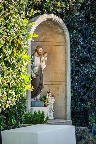 Statua di Gesù con bambino in nicchia di pietra tra arbusti verdi.