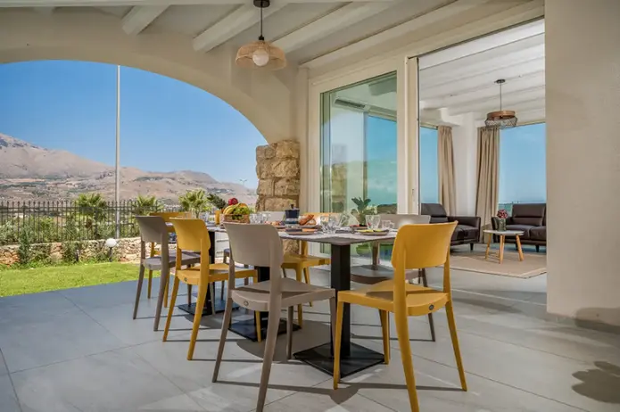 Patio esterno con tavolo da pranzo moderno, sedie colorate, vista mare e montagna.