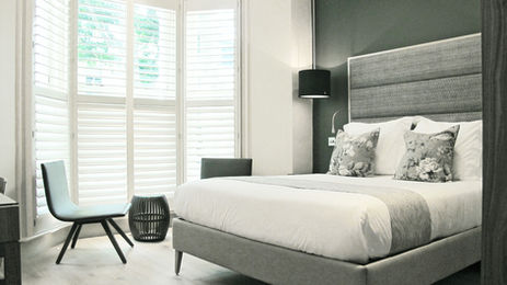 Light-filled King-bed guest room with space-making bay window, with comfortable seating for two people, tall layered upholstered headboard, elegant lighting and wooden floors