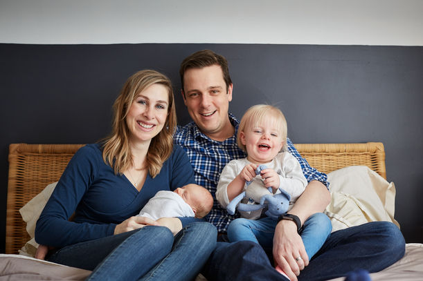 Family of 4 sitting together on a bed in their home smiling at camera