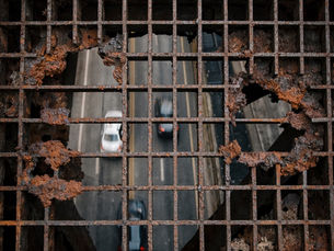 A top-down view through a rusted, industrial metal bridge grate with large jagged holes, looking down at cars driving on a busy highway below. The image represents the terrifying perspective of functional freeze and the precarious feeling of navigating a failing structure while under high stress.