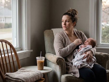 A woman sits in a worn armchair by a window, holding a sleeping newborn baby wrapped in a soft blanket. She has a thoughtful, slightly weary expression, gazing out the window into the soft daylight. Beside her, an iced coffee sits on a small white table. The scene captures the quiet, contemplative isolation often felt during the transition into motherhood. The lighting is soft and natural, emphasizing themes of maternal resilience, the "empty chair" of lost support systems, and the somatic experience of early parenthood.