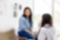 A woman wearing a blue button-up shirt and jeans is listening to a woman doctor in a comfortable exam room