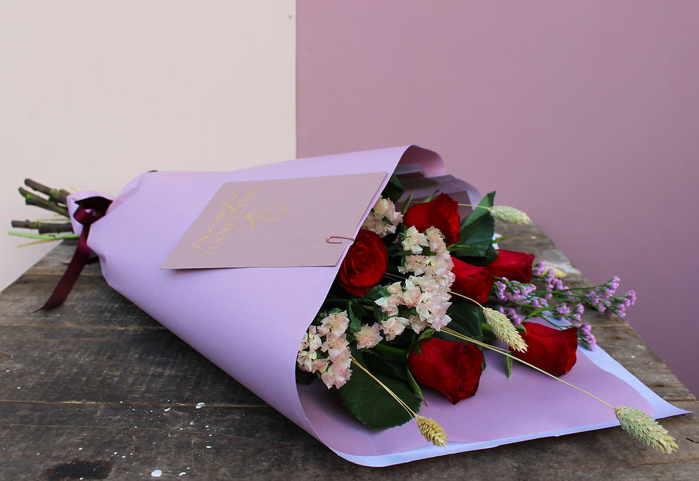 A bouquet of red roses and dried flowers wrapped in lilac paper with 'Wild About Flowers' branding, against pink and cream
