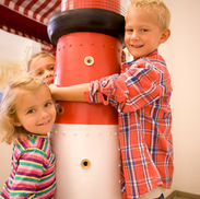 Drei lachende Kinder spielen um roten Leuchtturm-Spielzeug im Innenbereich.