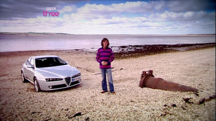 Top Gear Series 10 Episode 6 - James May standing next to the Alfa Romeo 159 at the base of the Humber Bridge on a pebble and sandy bank of the Humber River introducing the upcoming challenge.