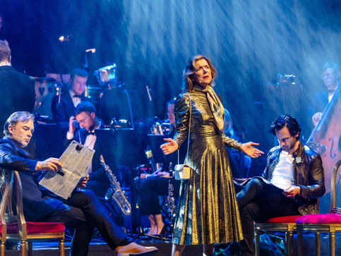 a woman in a gold dress stands on a stage in front of an orchestra