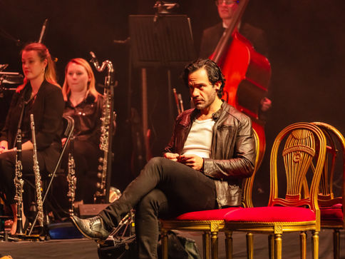a man in a leather jacket sits on a red chair in front of an orchestra