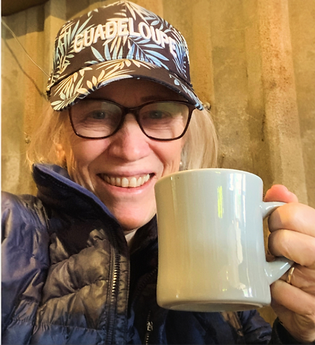 Robin Holland smiling and holding a coffee mug, wearing a “Guadeloupe” cap — casual, friendly portrait.