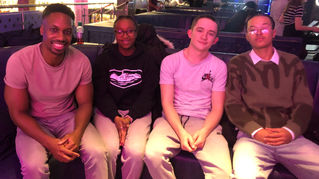 Four people seated on a blue couch, smiling. One wears a pink shirt, others in casual clothes. Warm lighting, relaxed atmosphere.