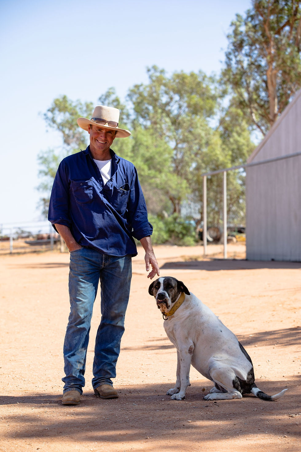 A man in a blue shirt and hat stands smiling beside a large dog on a sunny, dirt path with trees in the background.