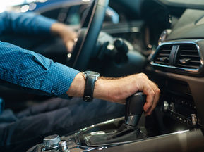 A person in a blue patterned shirt shifts a automatic car's gear in a modern interior. The dashboard is sleek, with a focus on precision and control.
