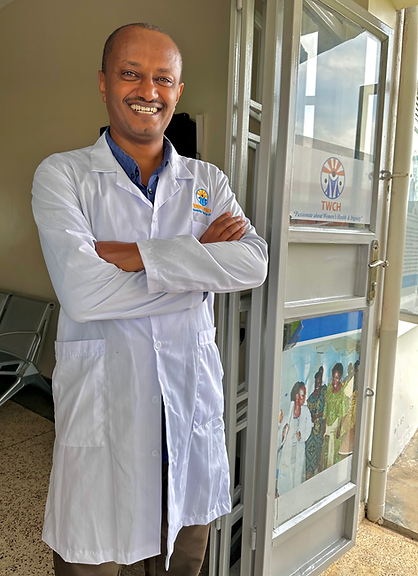 Man in white coat smiling, standing in the doorway at Terrewode Women's Community Hospital with a sign that reads"Passionate about Women's Health & Dignity."