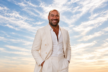 Niko-Helios in an ivory suit against a soft, natural cloud sky