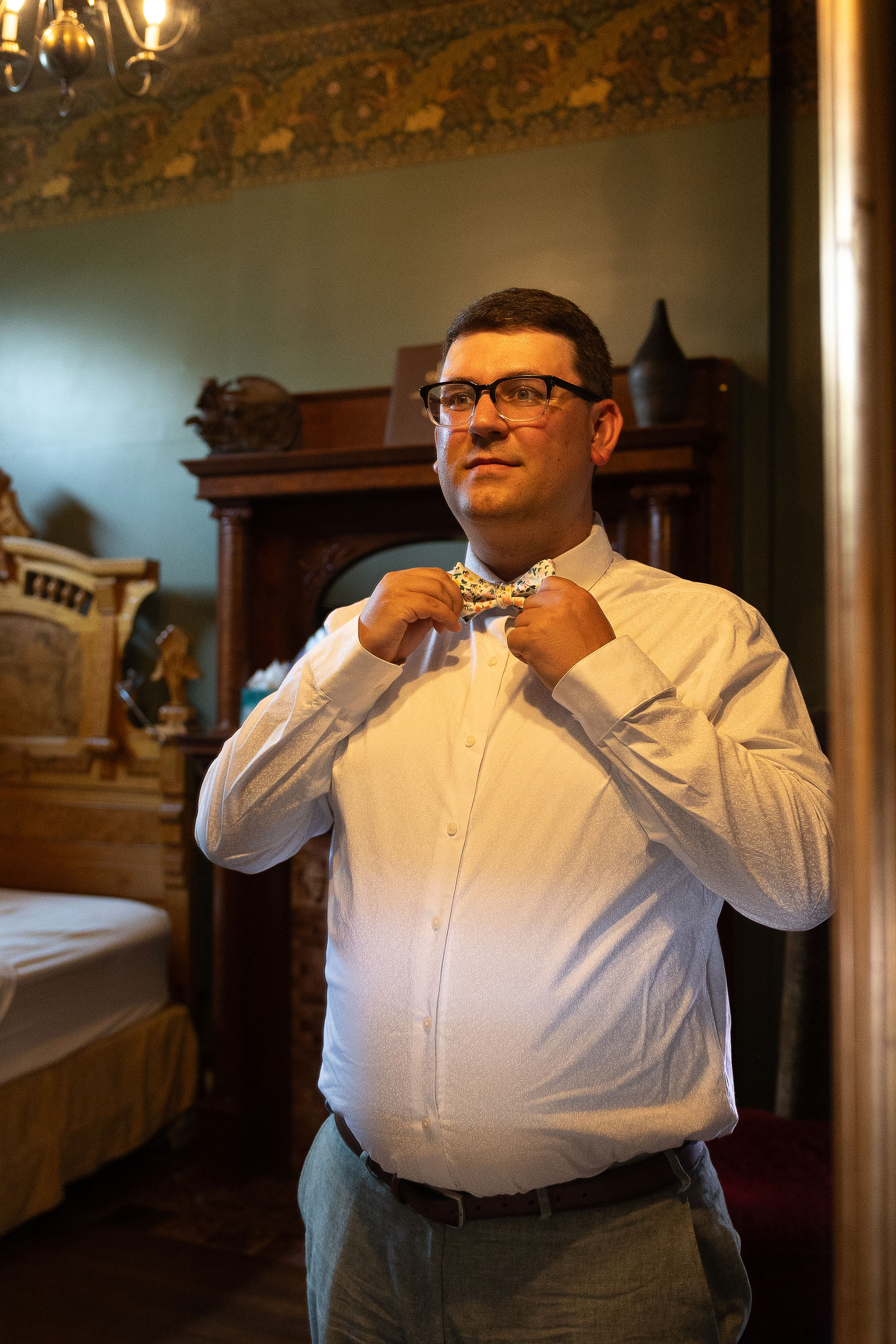 A groom adjusts his bow tie in front of a mirror.