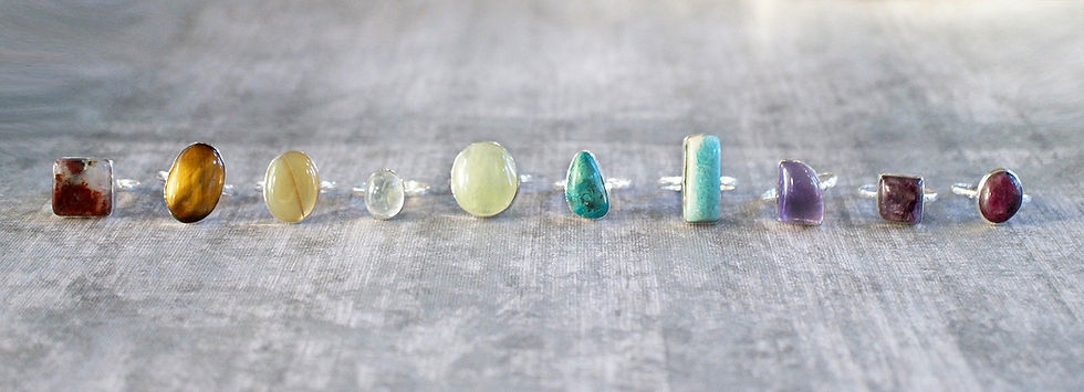 Nine gemstone rings on a textured grey surface, featuring various shapes and colors like brown, yellow, green, turquoise, and purple.