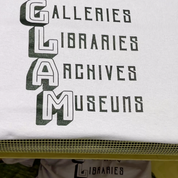 White shirt reads QUEER GLAM, Galleries, Libraries, Archives, Museums, on a dryer, screen printing t shirts with bold block lettering. At Wow! Custom Tees