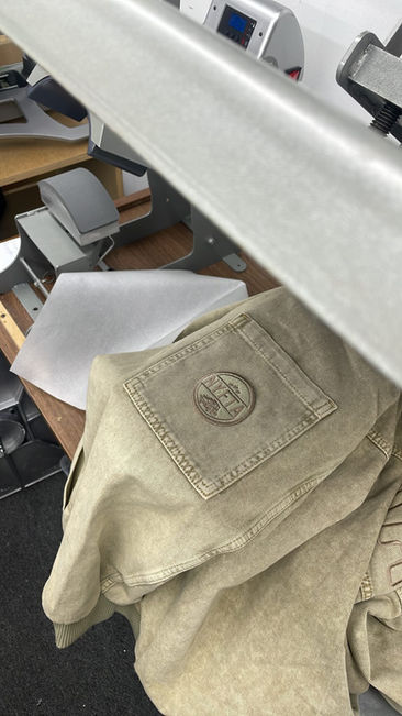 Light-tan embroidered denim jacket on a heat press with circular pocket patch; custom denim jacket being finished in a workshop. At Wow! Custom Tees