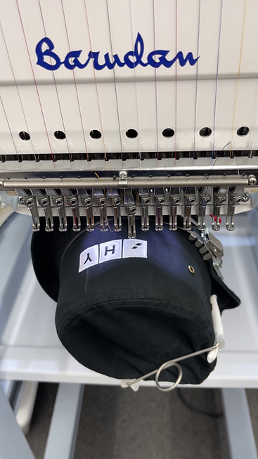 Close-up of an embroidery machine stitching a minimalist white icon trio onto a black embroidered bucket hat, under the needles. At Wow! Custom Tees.
