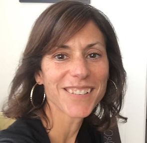 Headshot of Kristin Land. A woman with dark shoulder-length hair and large silver hoop earrings smiles warmly in a close-up photo against a light indoor background, wearing a dark top.