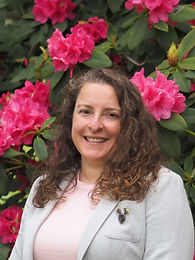 Headshot of Melissa Aguilar. A woman with dark curly hair and dark-rimmed glasses smiles warmly, wearing a black Puente-branded jacket and colorful beaded earrings, photographed outdoors against a blurred foliage background.