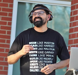 Headshot of Roberto Rubalcaba. A man with dark hair and a beard smiles while wearing over-ear headphones and a headband. His black t-shirt reads "Dream Like Martin, Read Like Harriet, Fight Like Malcolm, Write Like Garvey, Think Like Maya, Build Like Madam CJ, Speak Like Frederick, Educate Like W.E.B., Believe Like..., Challenge Like..." He stands outdoors in front of a brick building with a laptop visible in the foreground.