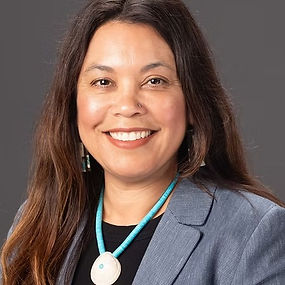 Headshot of Maria Figueroa, Training Coordinator. A woman with long dark hair smiles confidently, wearing a gray blazer and a turquoise beaded necklace with matching earrings, against a neutral gray background.