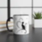 White mug with black interior and handle, cat silhouette with hearts, placed on desk with plant and books.