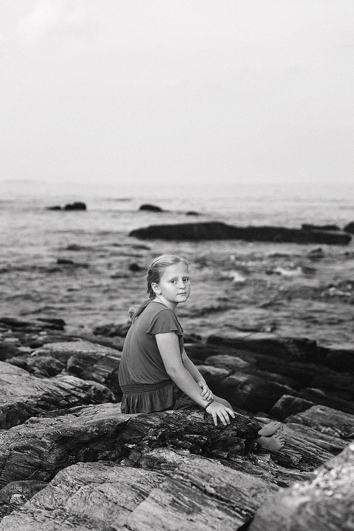Black and white image of young girl on the rocks in Maine with water in the background