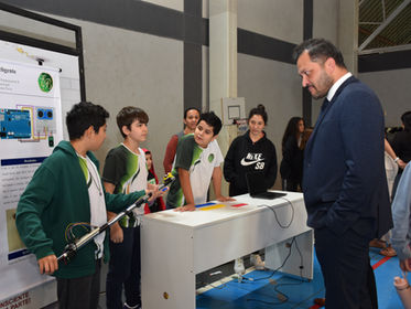 Grupo de estudantes e homem observam projetos em exposição Início Escola Erasmo Braga