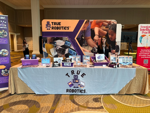 Exhibit booth with "True Robotics" banner, showcasing robotics kits and materials. A smiling person stands behind a table with promotional items.