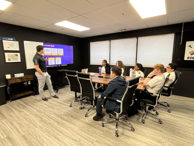 Small group of students seated around a conference table in a meeting room while an instructor stands beside a screen labeled “Startup Phase,” pointing to a presentation slide; certificates and a logo are displayed on the walls.
