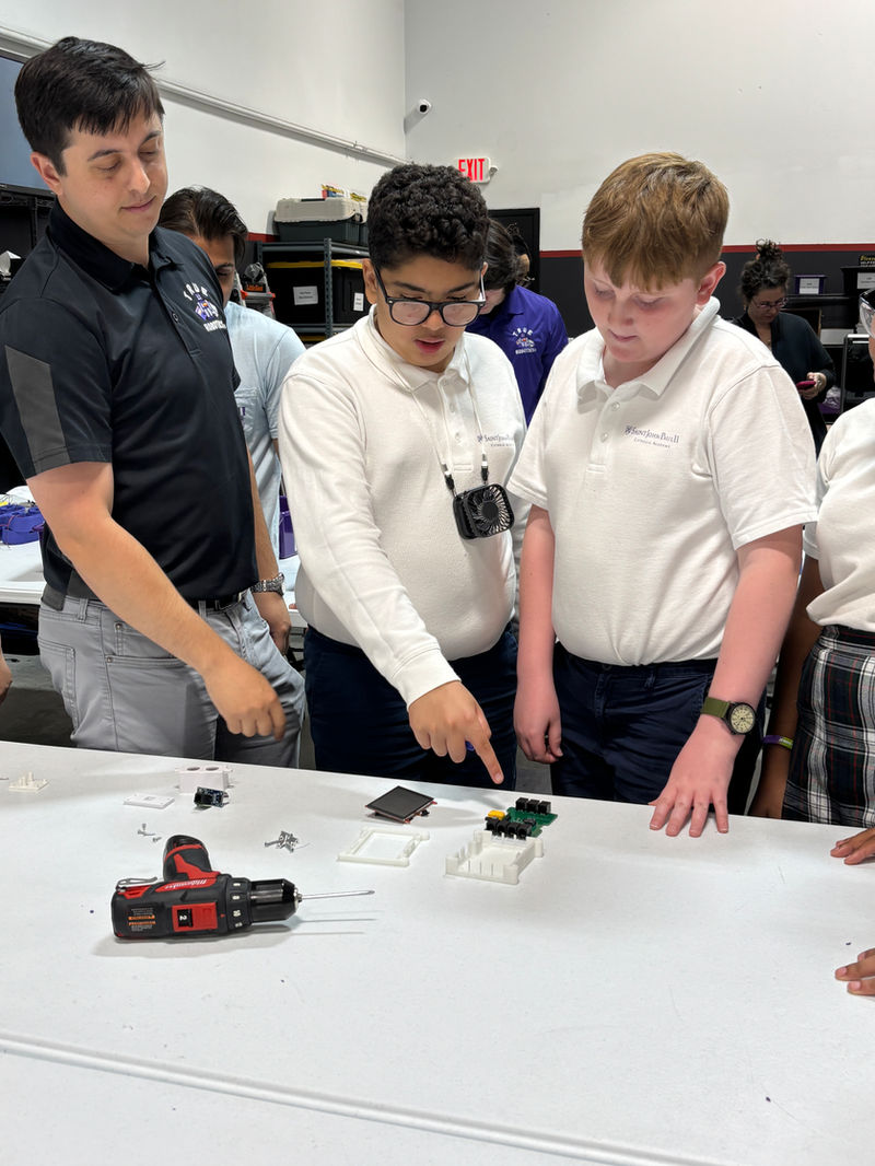 Students and an instructor gather around a worktable in a robotics lab, examining electronic components and parts spread out on the surface, including a small circuit board, screws, and a cordless drill.