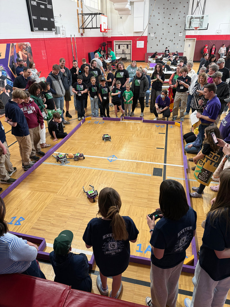 Students, educators, and families gathered around a robotics competition field in a gym, watching small robots navigate a marked course while teams operate controllers and observe closely.