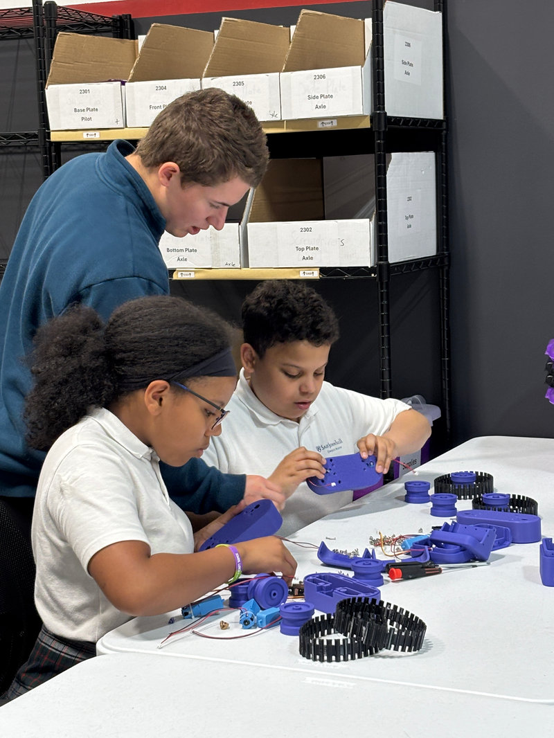 Students work at a table assembling purple robotic components while an instructor leans over to guide them; shelves behind them hold labeled boxes of parts such as “Base Plate,” “Front Plate,” and “Side Plate Axle.”