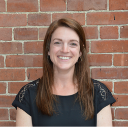 Portrait of a smiling woman with long brown hair wearing a black top, standing in front of a red brick wall.