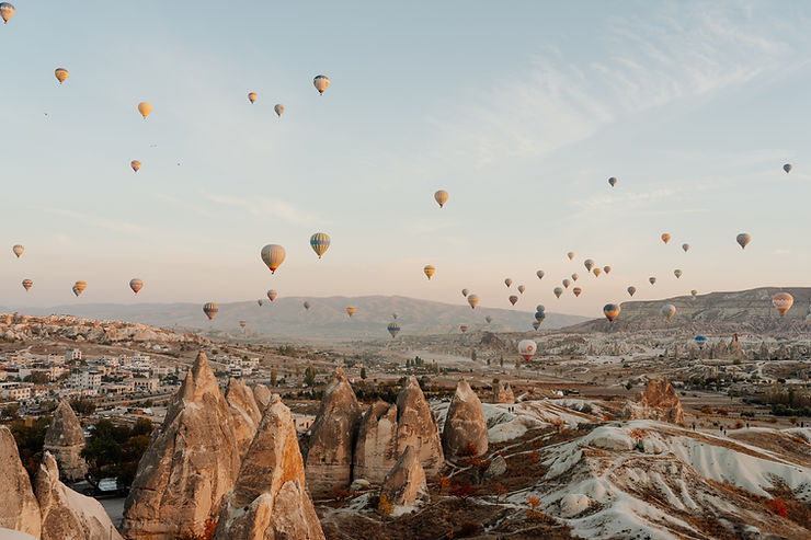 Hot air balloons drift over Cappadocia’s rocky valleys and town at sunrise, with soft pastel sky and distant hills.