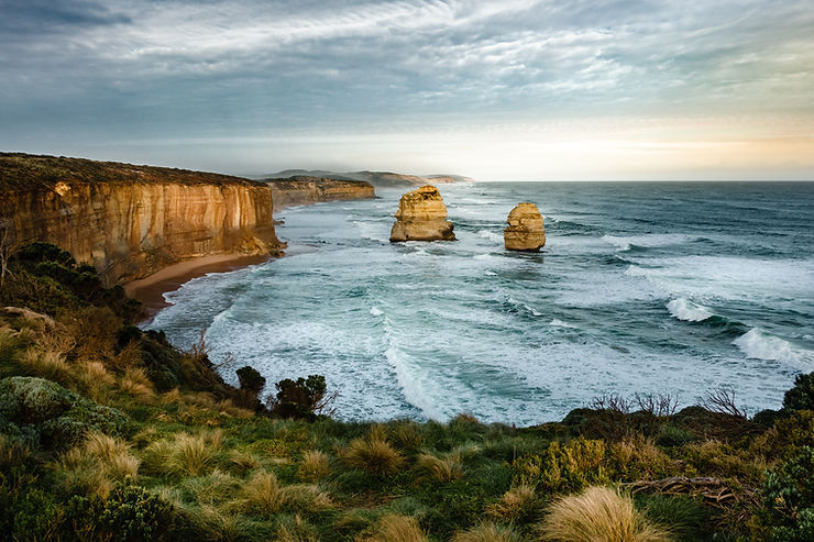 Windy ocean bay with two sea stacks, golden cliffs, grassy foreground, and a cloudy sky.