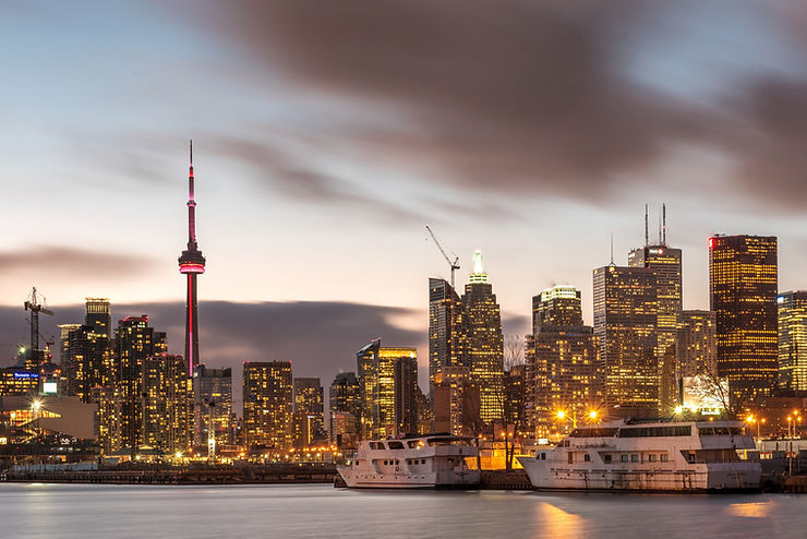 Toronto skyline at dusk with the CN Tower glowing red, lit high-rises, boats on the water, and a cloudy sky.