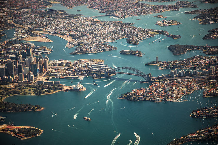 Aerial view of Sydney Harbour with the Harbour Bridge, Opera House, city skyline, and boats crossing blue water.