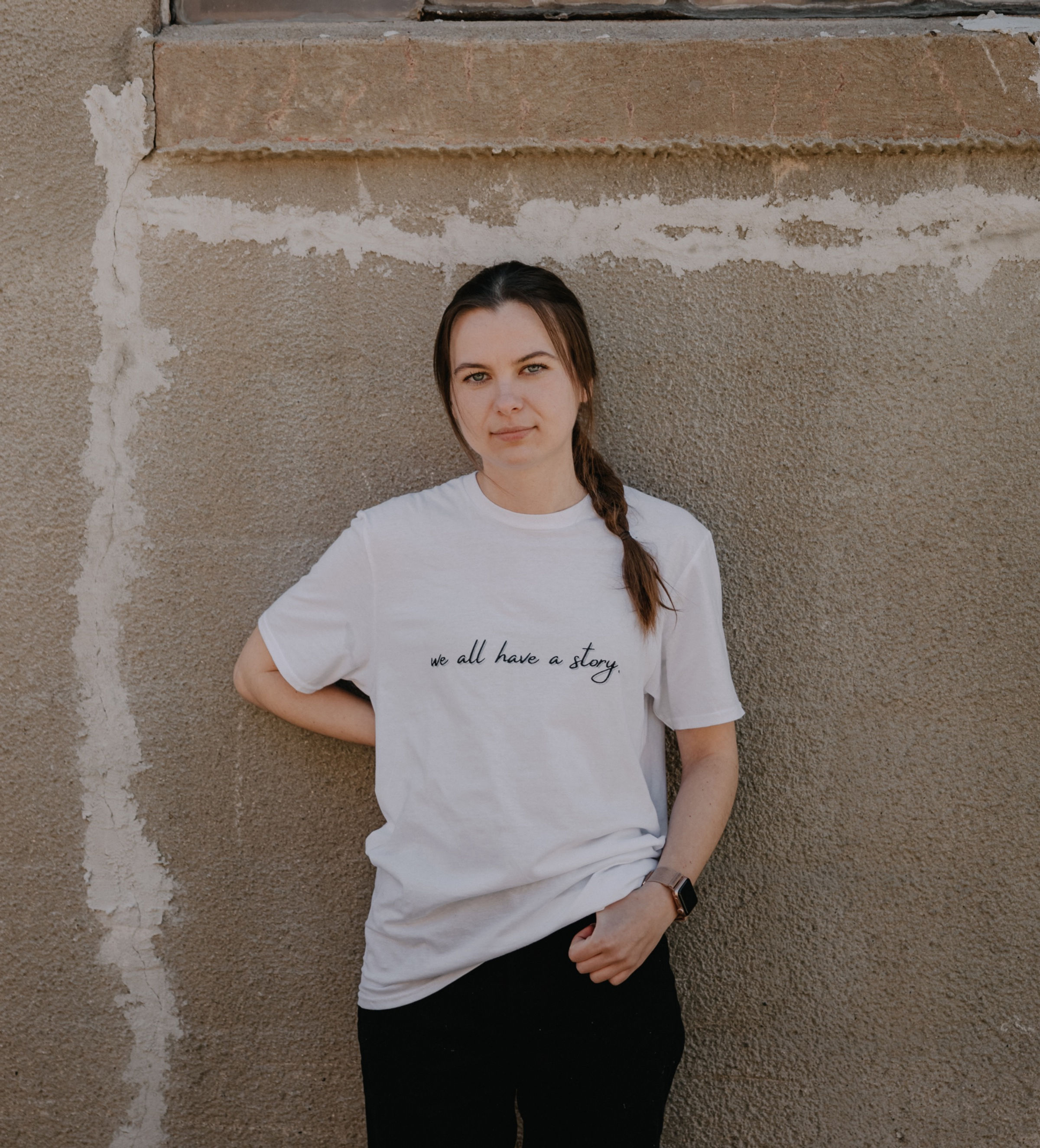 White Cotton T-Shirt that says, "We All Have a Story" in small, cursive letters across the chest area.